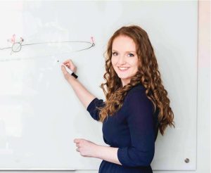 Juliette Becker looks back at the camera while writing on a. whiteboard. She is smiling, wearing a blue top.